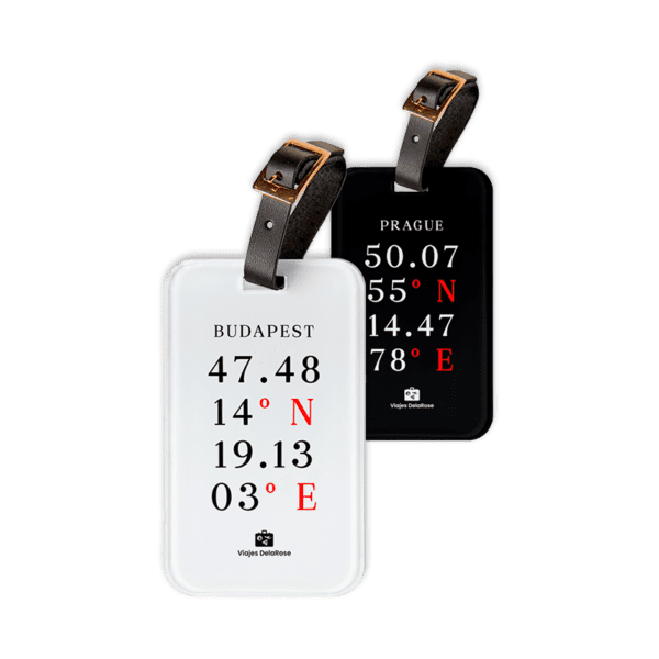 Dos etiquetas de equipaje rectangulares, una blanca y otra negra, con coordenadas de ciudades en tipografía elegante. Ambas tienen una correa de cuero con hebilla dorada para sujetarlas a la maleta.